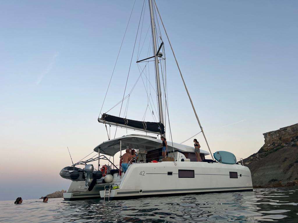 Bowie Lagoon 42 catamaran anchored near Malta's coastline at sunset with guests swimming